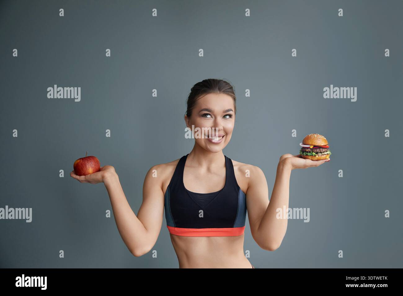 Eine Frau steht mit einem Apfel in der linken Hand und einem Burger in der rechten und lächelt selbstbewusst. Sie trägt ein sportliches Outfit und ist gegen einen einfachen bac gestellt Stockfoto