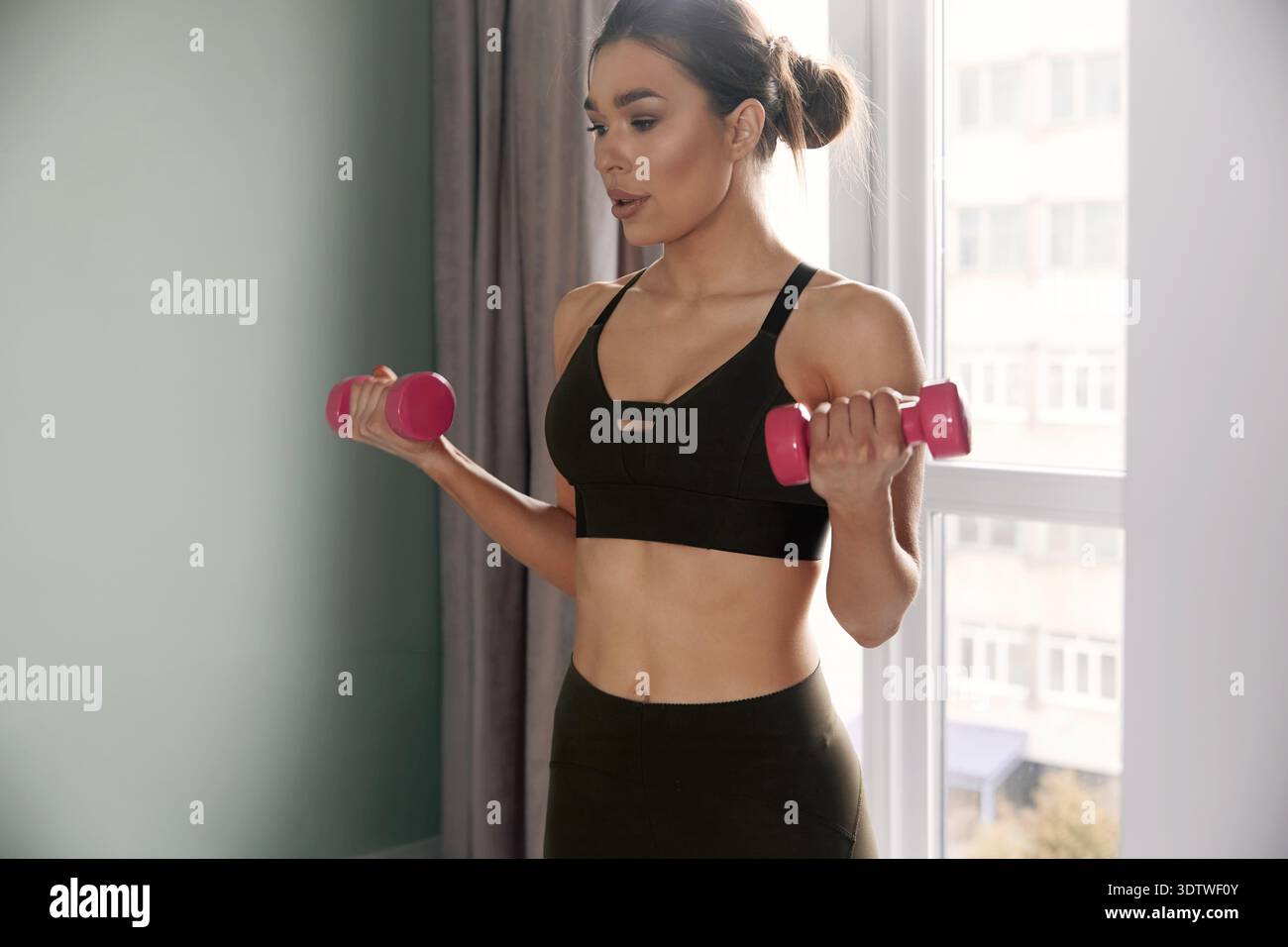 Eine Frau arbeitet zu Hause und hebt rosa Kurzhanteln nahe dem Fenster. Das Nachmittagslicht leuchtet auf sie und den Raum. Stockfoto
