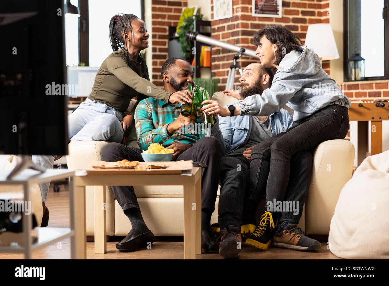 Fröhliche multiethnische Freunde feiern gemeinsam auf der Couch, heben Bierflaschen auf und teilen sich Snacks im gemütlichen Wohnzimmer mit Ziegelwänden. Junge Menschen, die sich gerne in ungezwungener Atmosphäre aufhalten und zu Hause gesellig sind. Stockfoto