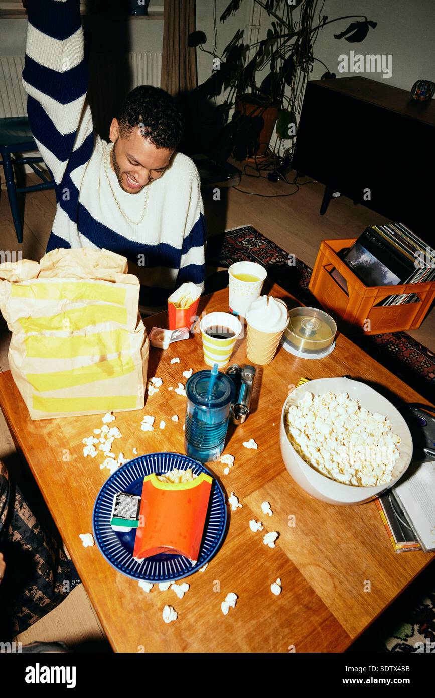 Fröhlicher Mann mit Fastfood auf dem Tisch zu Hause Stockfoto