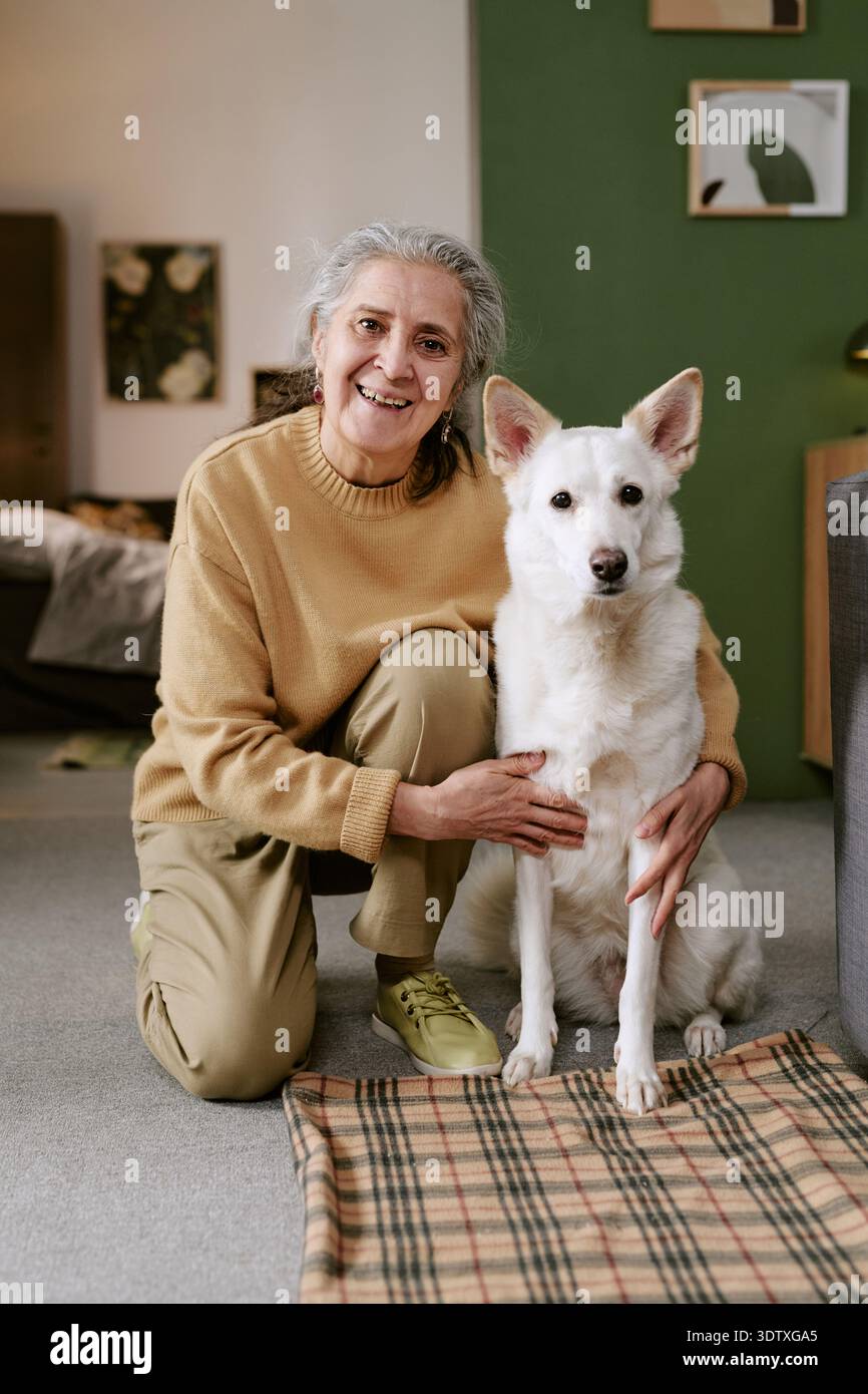 Porträt einer älteren kaukasischen Frau, die lächelt, während sie einen Hund im Haus streichelt Stockfoto