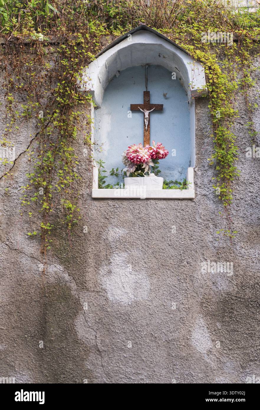 Ein katholischer Schrein am Straßenrand in einer blau bemalten bogenförmigen Nische auf einer verwitterten Stuckwand mit einem Kruzifix und rosa Dahlienblüten in einer weißen Vase Stockfoto