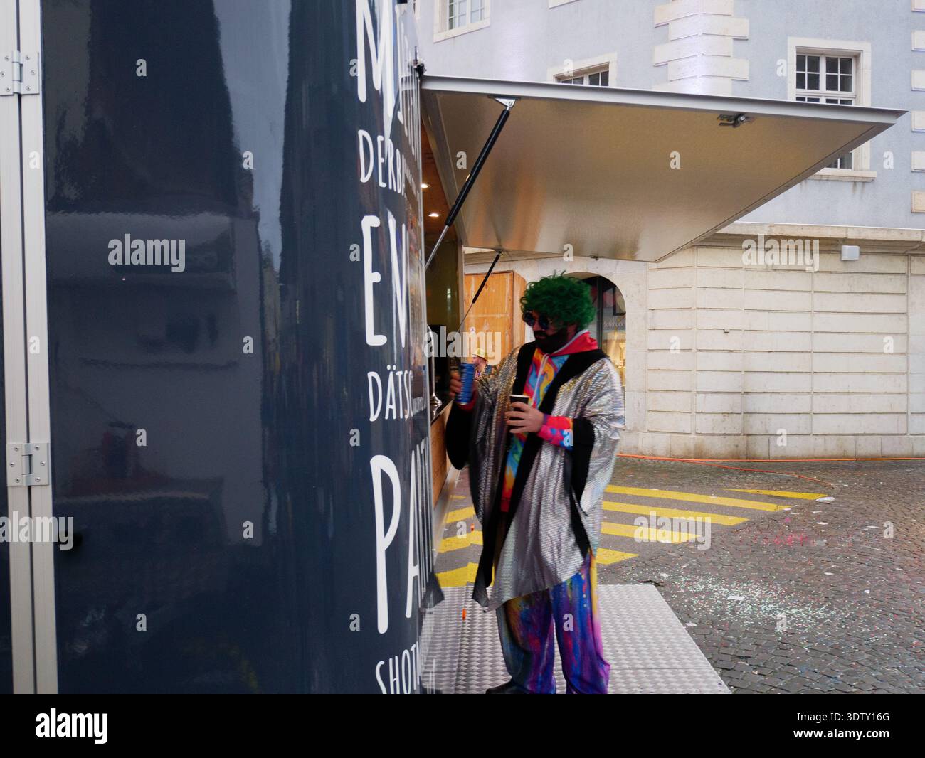 Langenthal, Bern, Schweiz. Februar 2026. Fasnacht Karneval in der Stadt. Ein Clown in farbenfroher Kleidung steht neben einem Food Truck in einer städtischen Umgebung Stockfoto