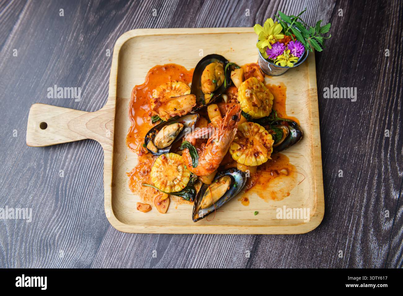 Blick von oben auf scharfe, gebratene Meeresfrüchte mit Garnelen, Muscheln und Babymais, elegant auf einem Holzteller mit zarter Blumengarnitur serviert. Stockfoto