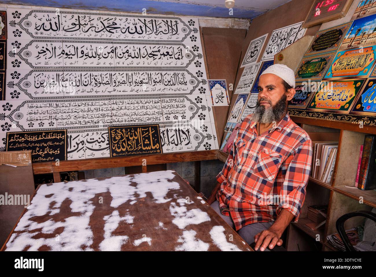 Ein Mann in einem karierten Hemd sitzt in einem kleinen Geschäft, umgeben von dekorativen Fliesen und Tafeln mit arabischer Kalligraphie in Alt-Delhi. Stockfoto