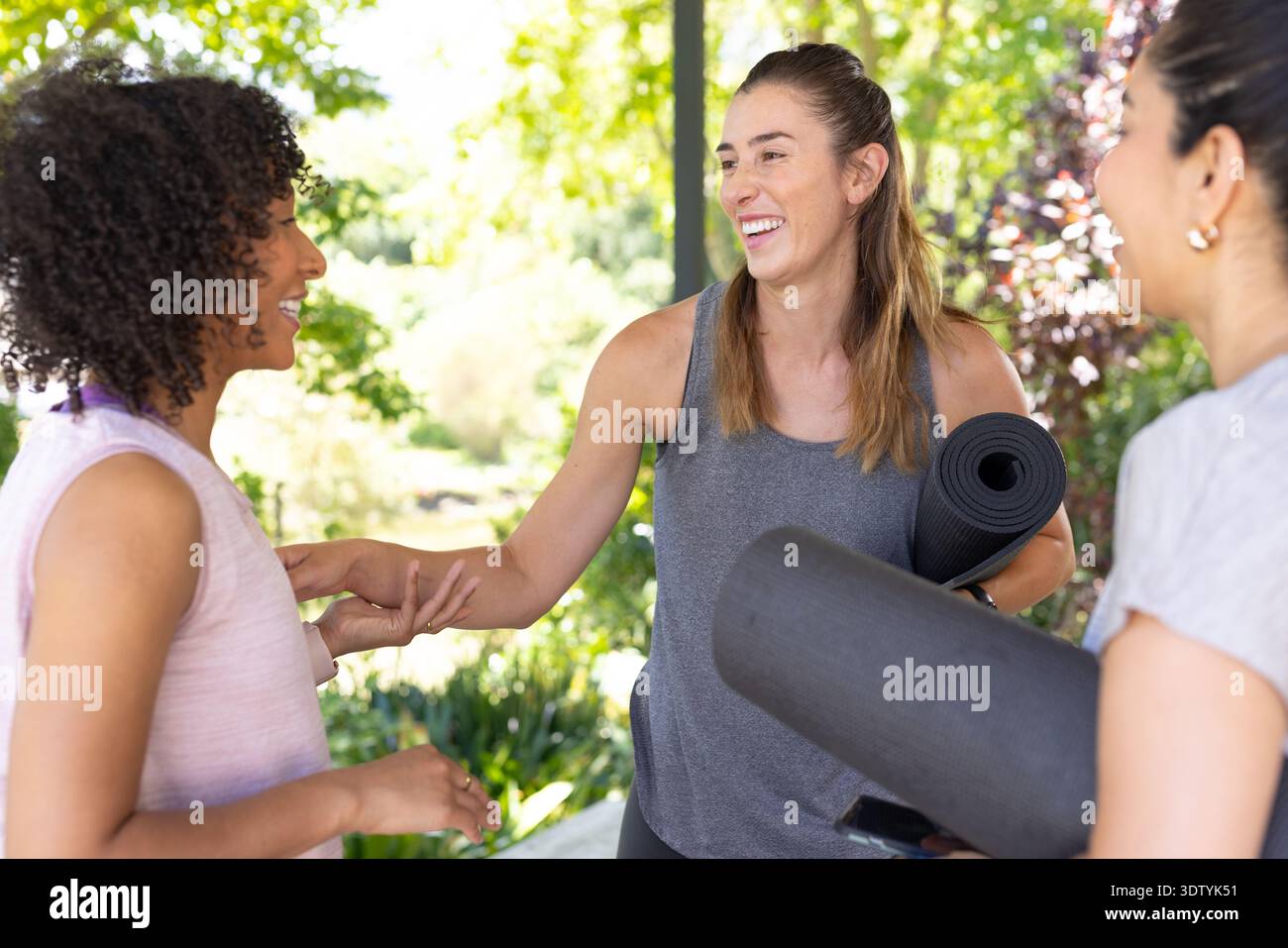 Verschiedene Freundinnen lächeln und unterhalten sich in Aktivkleidung mit schwarzen Yogamatten und Telefon im Park Stockfoto