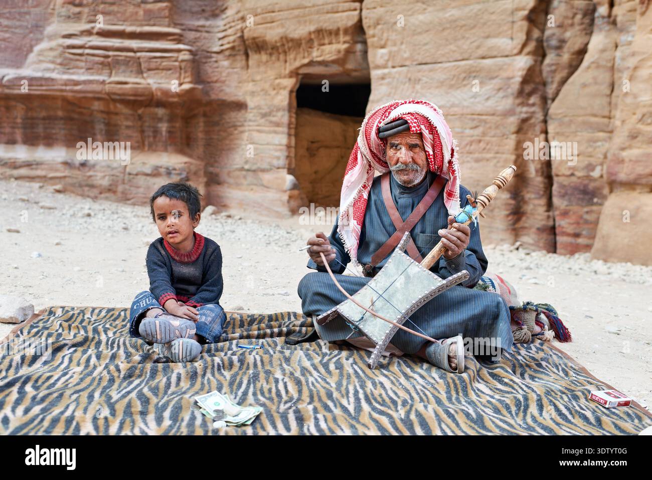 Ein Mann spielt ein traditionelles Musikinstrument in Petra, während ein Kind in der Nähe sitzt. Sie sind umgeben von alten Felsen und Geschichte. Stockfoto