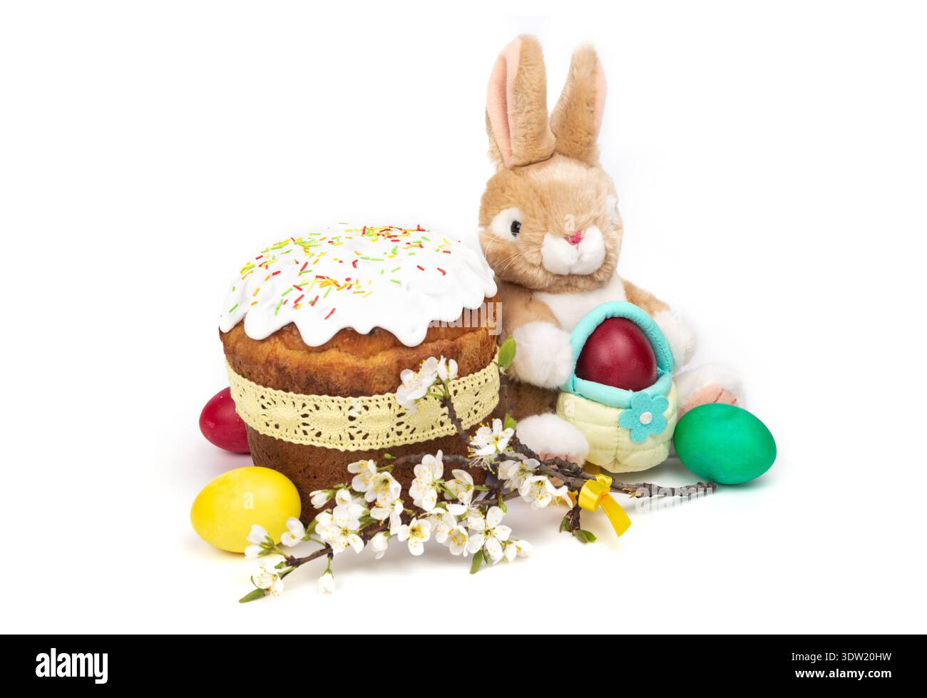 Ein Hasenspielzeug sitzt neben einem mit Glasur verzierten Kuchen, umgeben von bunten Eiern und Blumen, die das Frühlingsfest markieren. Stockfoto