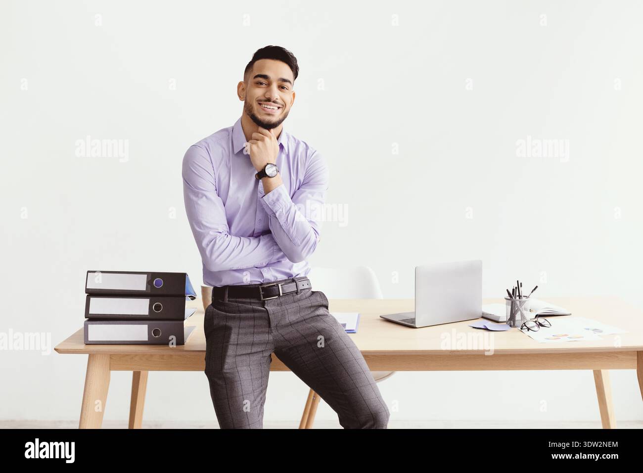 Professioneller Mann lächelt am Schreibtisch, während er in einem modernen Büro bei hellem Tageslicht arbeitet Stockfoto