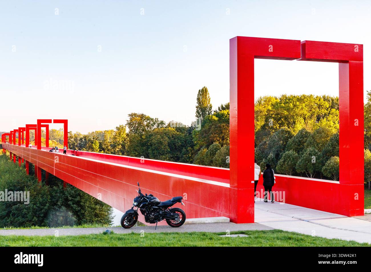 Eine markante rote Fußgängerbrücke überspannt eine üppige grüne Landschaft mit modernem architektonischem Design. Stockfoto