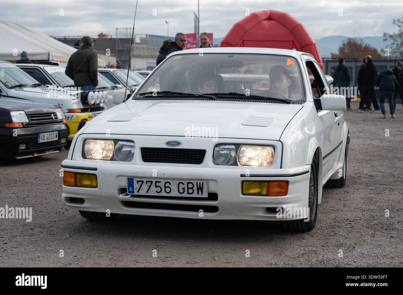 Vorderansicht eines weißen Ford Sierra RS Cosworth klassischen Sportwagens mit beleuchteten Scheinwerfern, der durch eine Outdoor-Veranstaltung fährt. Stockfoto