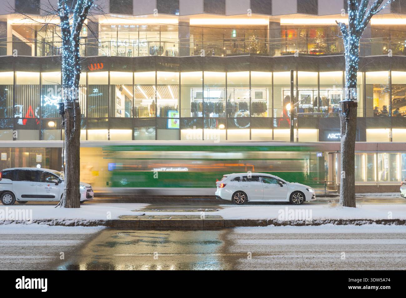 Lange Zeit im verschneiten Stadtzentrum von Sapporo, Hokkaido, Japan, ist ein verschwommener Bus an geparkten Autos und neonbeleuchteten Einkaufszentren vorbeigefahren. Stockfoto