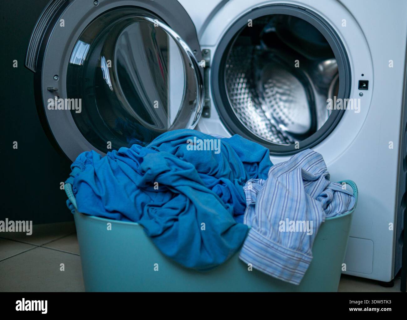 Korb gefüllt mit schmutziger blauer und gestreifter Kleidung vor einer offenen Waschmaschine. Innenraum-Szene mit Vorbereitung auf die Wäsche. Stockfoto