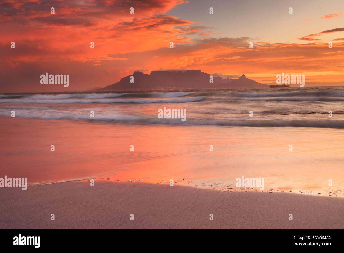 Blick auf den legendären Tafelberg, der sich vor einem feurigen Sonnenunterganghimmel befindet, Wellen, die sanft das Sandstrand wäschen und eine ruhige Landschaft schaffen, Kapstadt Stockfoto