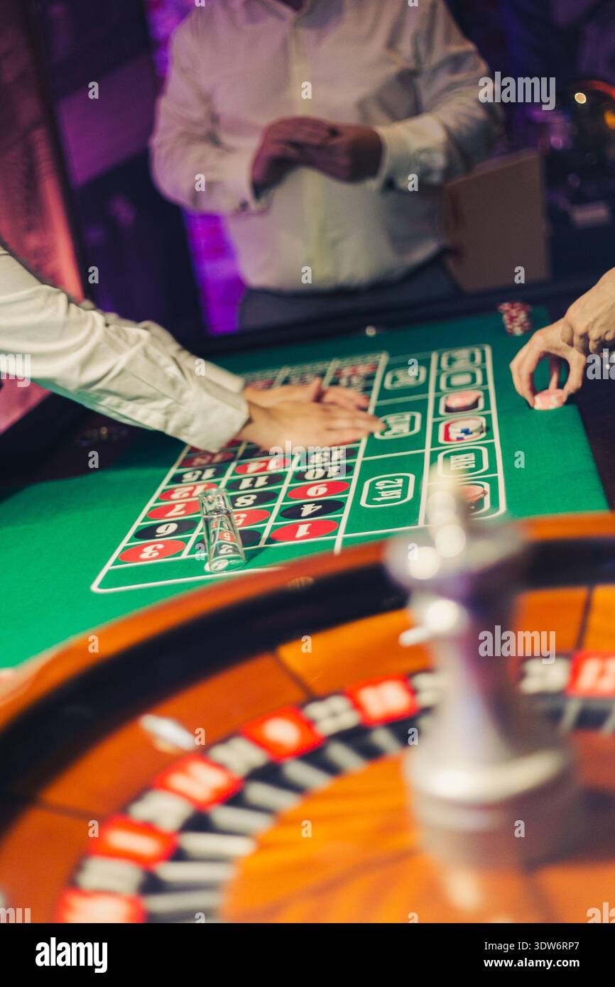 Eine dynamische Szene an einem Roulette-Tisch, in der Spieler Wetten auf dem grünen Filzlayout platzieren. Das Roulette-Rad ist im Vordergrund sichtbar. Stockfoto