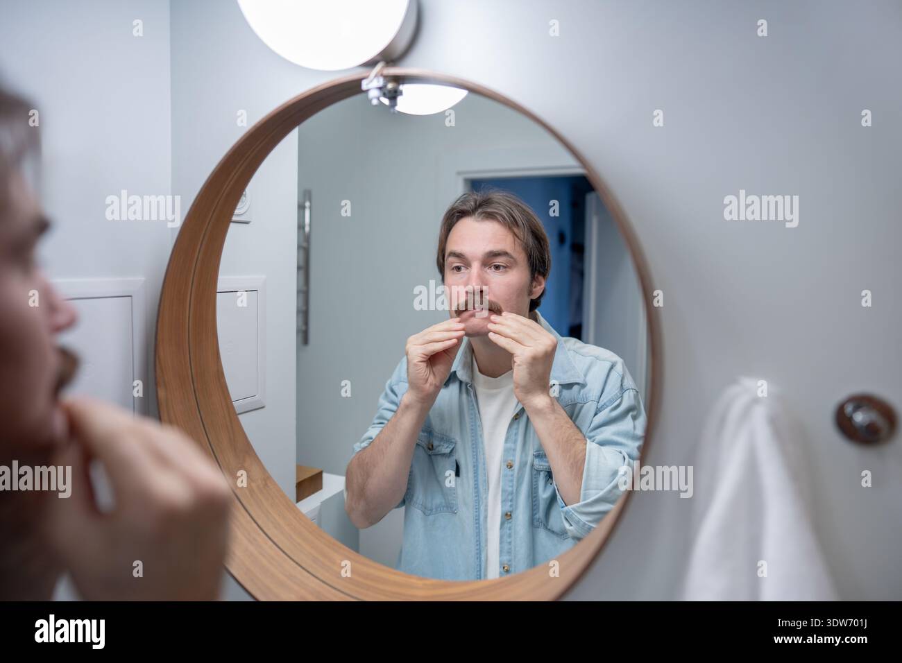 Junger Mann mit Schnurrbart schaut auf die Reflexion im Spiegel im Badezimmer. Hautpflege-Morgenroutine für Männer. Stockfoto