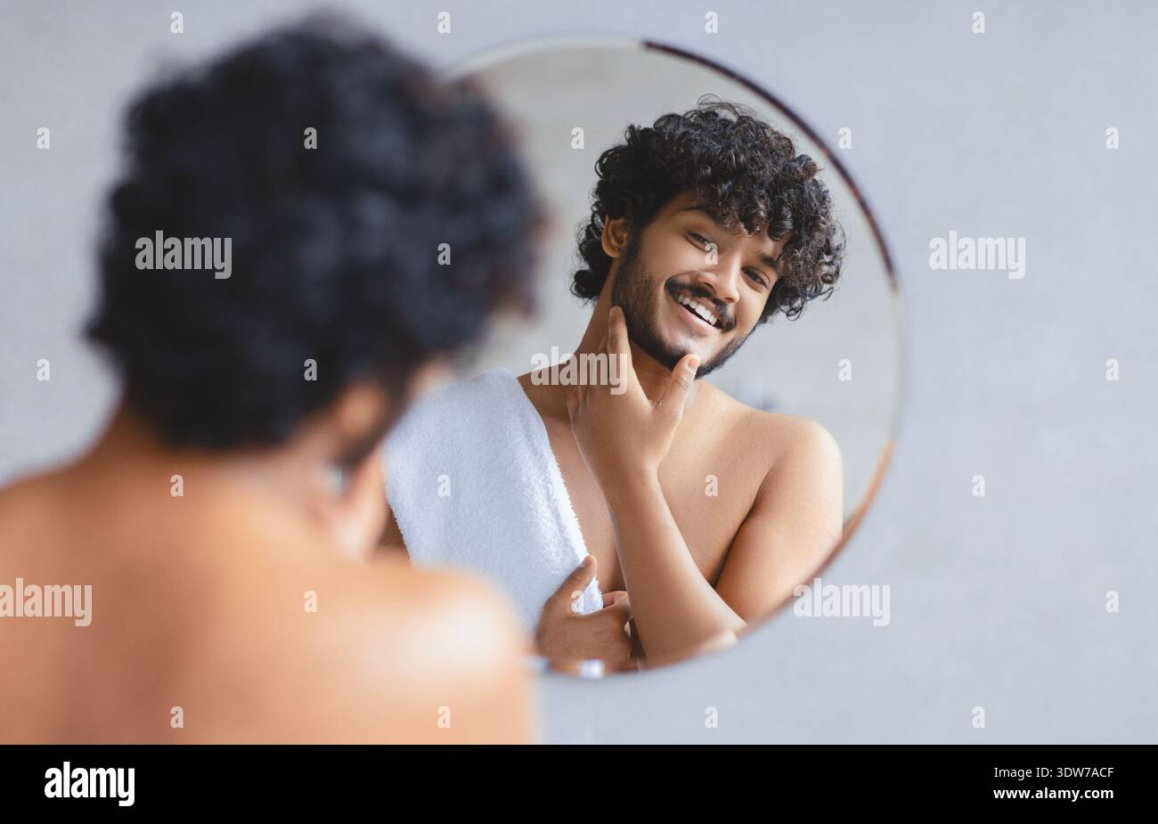 Junger Mann, der sich im Spiegel anfühlt und lächelt, während der Morgenroutine im Badezimmer Stockfoto