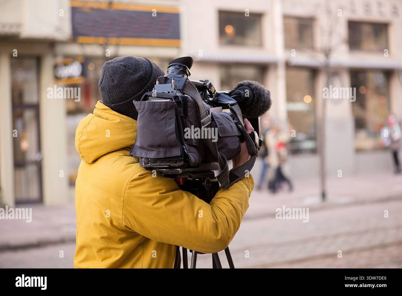Kameramann mittleren Alters, der eine gelbe Jacke trägt und eine professionelle Videokamera auf einem Stativ in einer städtischen Straßenumgebung bedient. Stockfoto