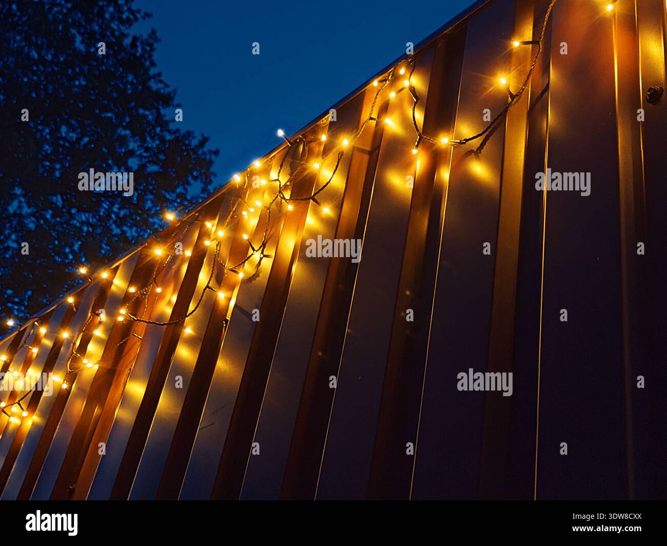 Warme gelbe Lichter hängen bei Dämmerung an einer Metallwand und leuchten sanft am tiefblauen Abendhimmel und schaffen eine gemütliche festliche Atmosphäre. Stockfoto