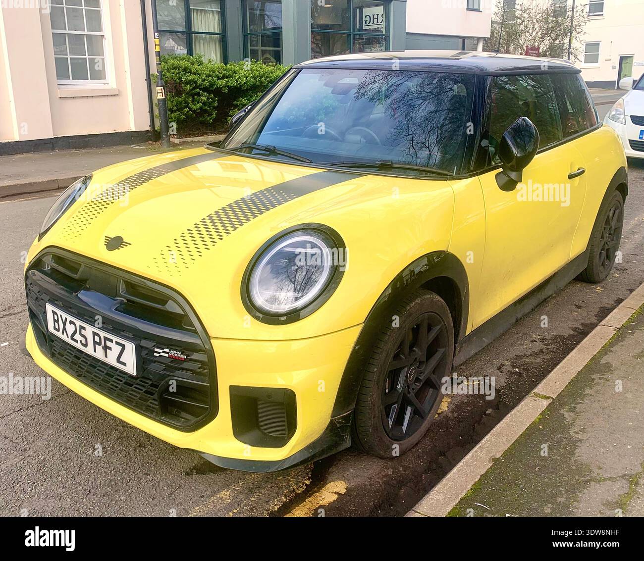 Ein leuchtend gelber Mini-cooper mit schwarzen Rennstreifen, der entlang einer ruhigen Wohnstraße geparkt wird, mit sportlichem Design und auffälligen Farben im Hintergrund - Smartphone-aufgenommenes Stockfoto