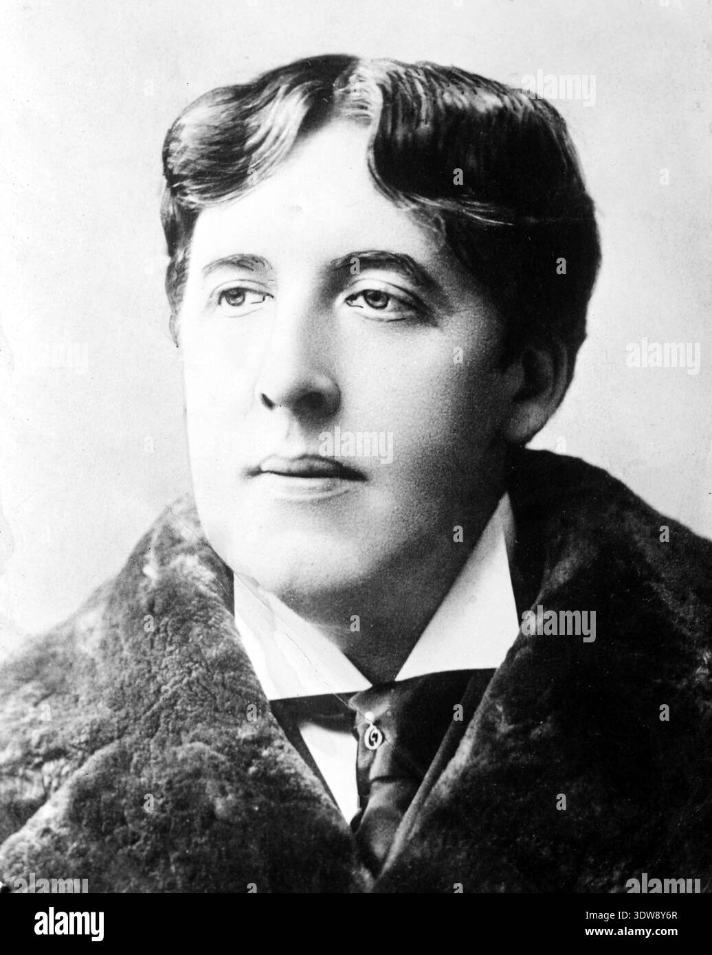 Dieses schwarz-weiße Porträt aus den 1880er Jahren zeigt Oscar Wilde, den bekannten irischen Dichter und Dramatiker, der mit einem ernsten Ausdruck zur Seite blickt. Er trägt einen Mantel mit Pelzkragen, ein weißes Hemd und eine dunkle Krawatte, die die Mode des späten 19. Jahrhunderts widerspiegelt. Stockfoto