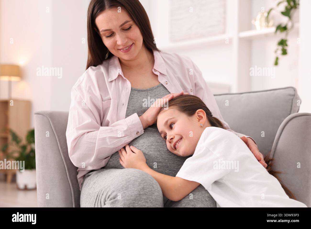 Schwangere Frau und ihre Tochter sitzen zu Hause auf einem Sessel Stockfoto