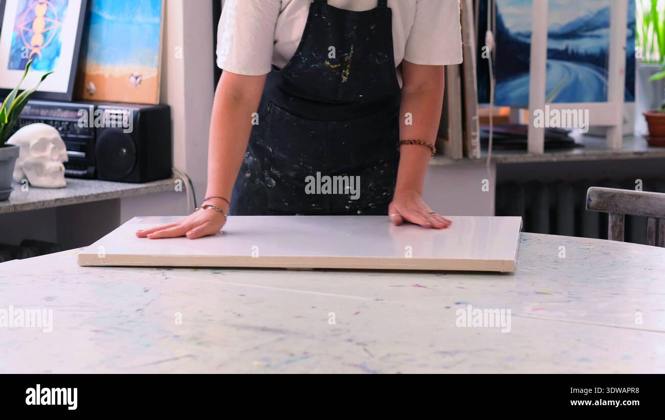 Die offene Studioszene zeigt eine Person, die eine große weiße Leinwand auf einem Tisch mit Farbspritzern positioniert, darunter Kunstwerke und Werkzeuge. Planung der Stockfoto