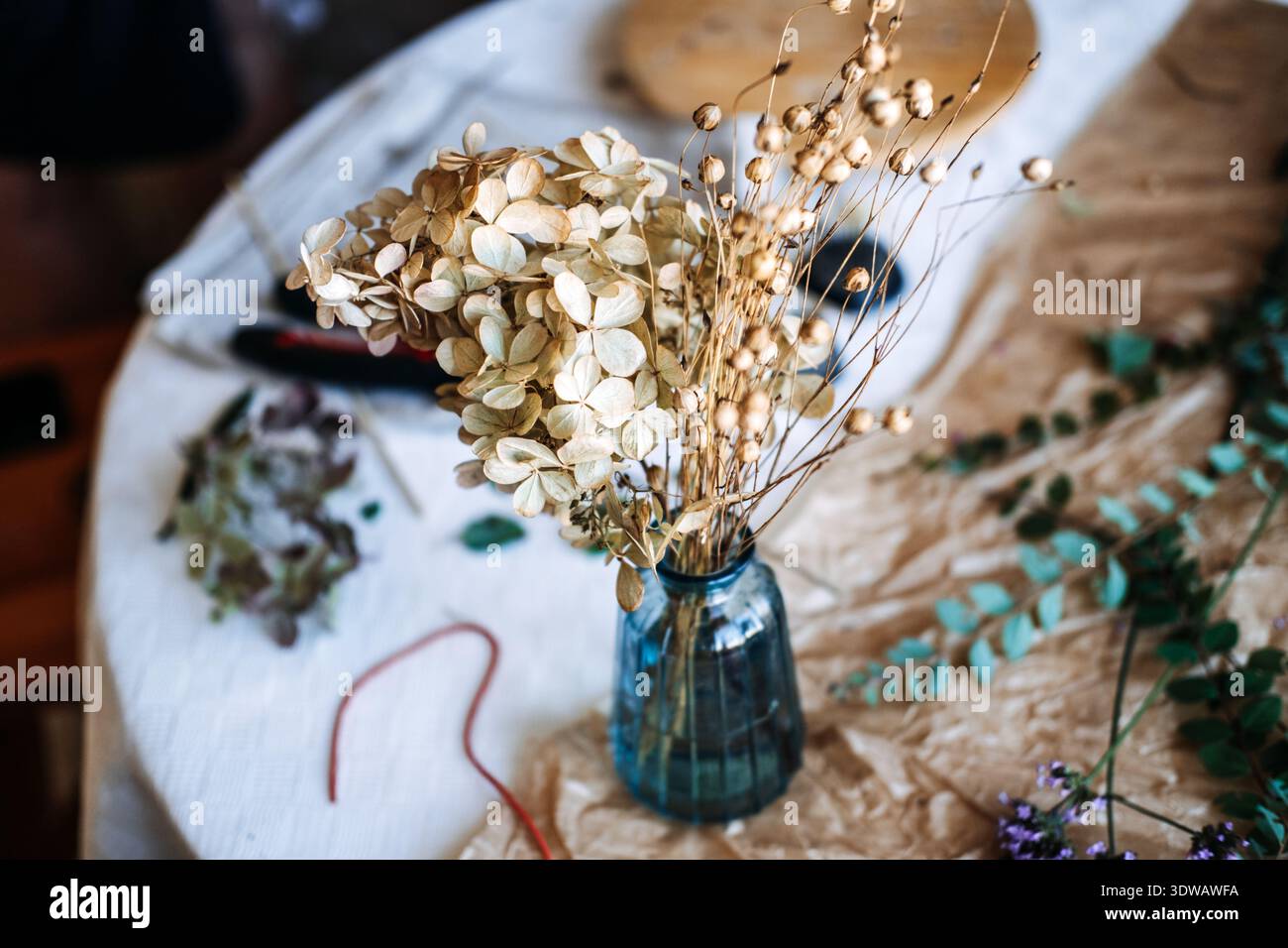 Getrocknete Hortensie und Samenkapseln stehen in einer kleinen blauen Glasvase auf dem Tisch mit botanischen Trümmern. Verfaulende florale Schönheit, trockene Blumenküche, Schönheit Stockfoto