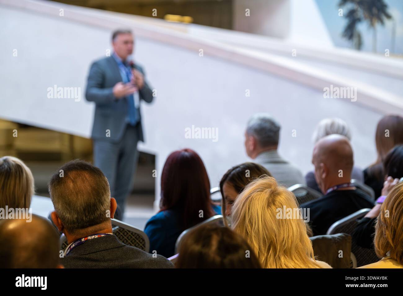 Speaker in Suit erklärt den verschiedenen Gruppen von Fachleuten im hellen Lobbybereich Ideen, die in modernem Veranstaltungsort mit Handgesen lauschen und dabei ansprechende Geschäftsbotschaften vermitteln. Stockfoto