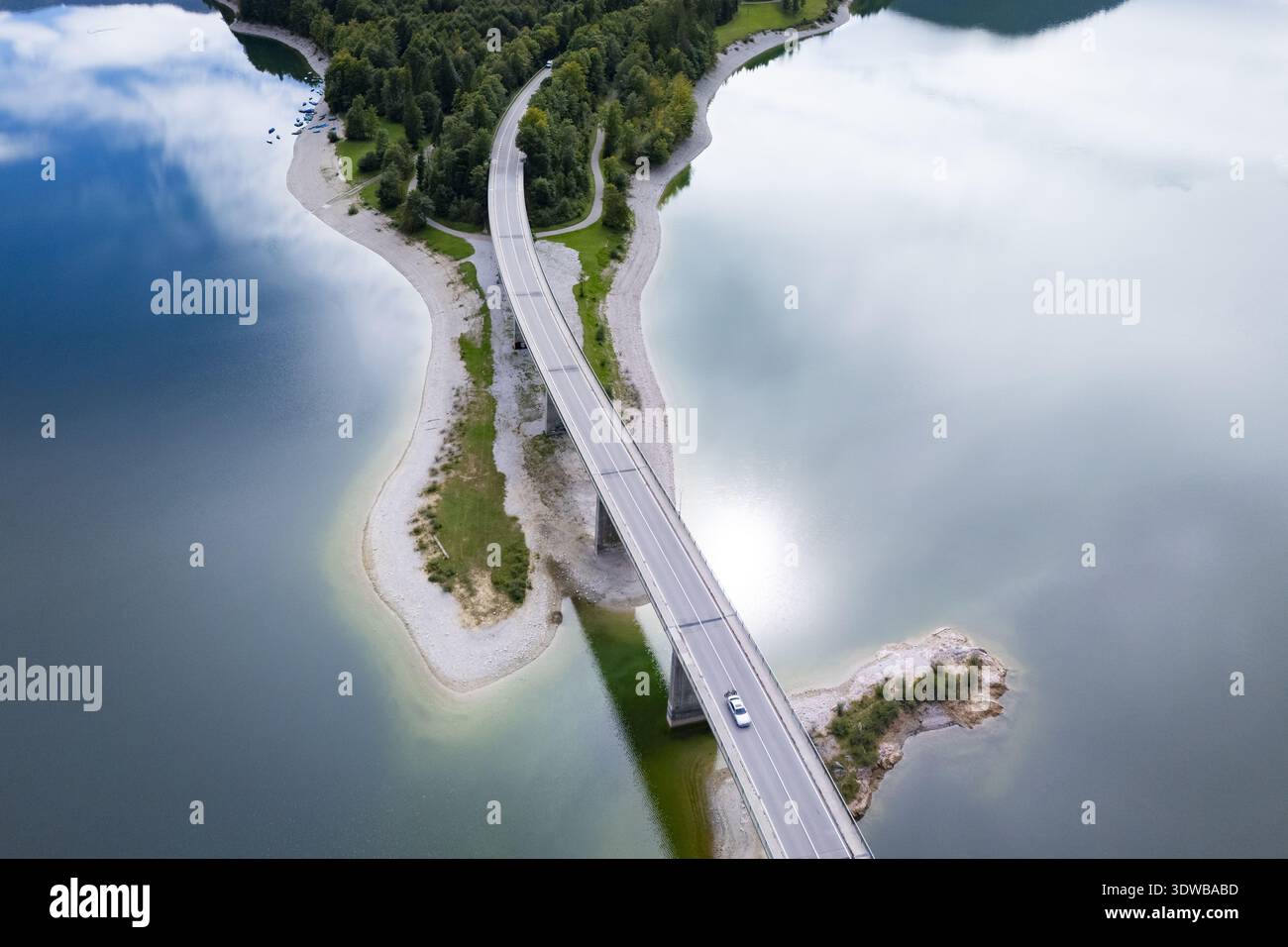 Aus der Vogelperspektive einer Brücke, die durch den ruhigen See führt und den Himmel spiegelt und üppige grüne Landmassen verbindet, Sylvensteinspeicher, Bayern, Deutschland. Stockfoto