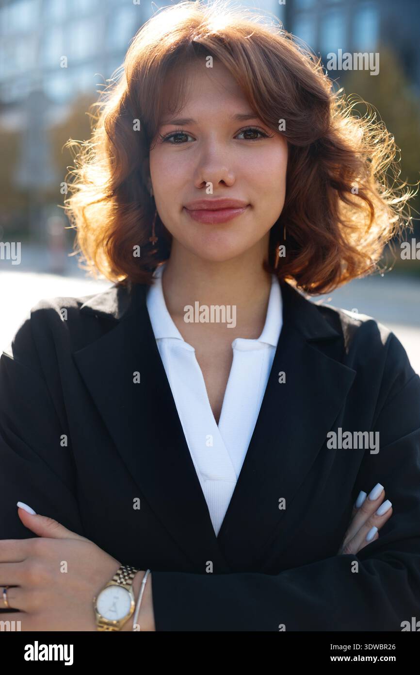 Selbstbewusste Geschäftsfrau mit gekreuzten Armen posiert in moderner städtischer Umgebung mit Führungseinstellung. Stockfoto