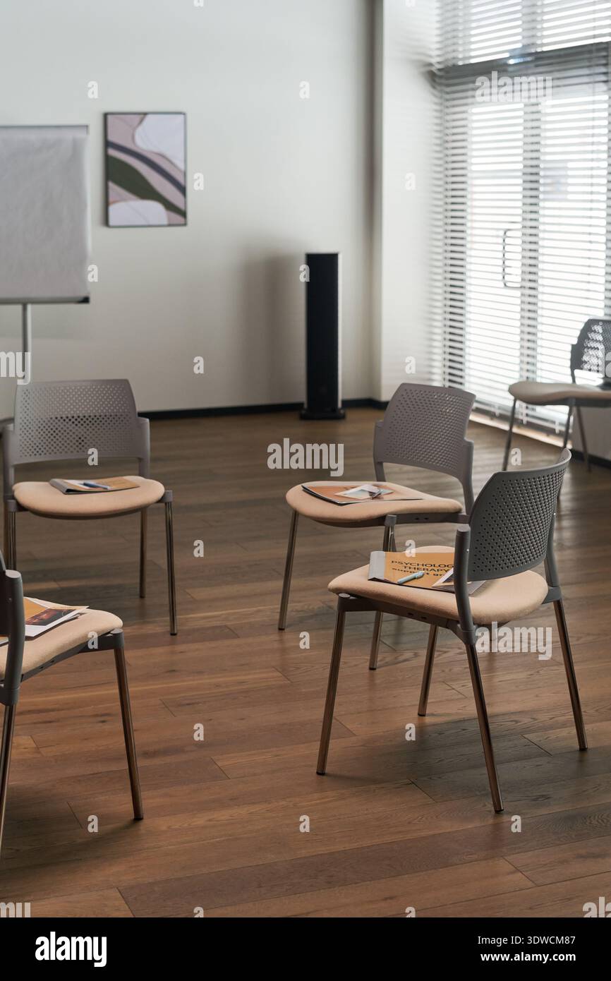Leerer, moderner Tagungsraum mit kreisförmigen Stühlen, Notizbüchern und Stiften auf den Sitzen, Holzboden und großen Fenstern mit Jalousien für natürliches Licht Stockfoto