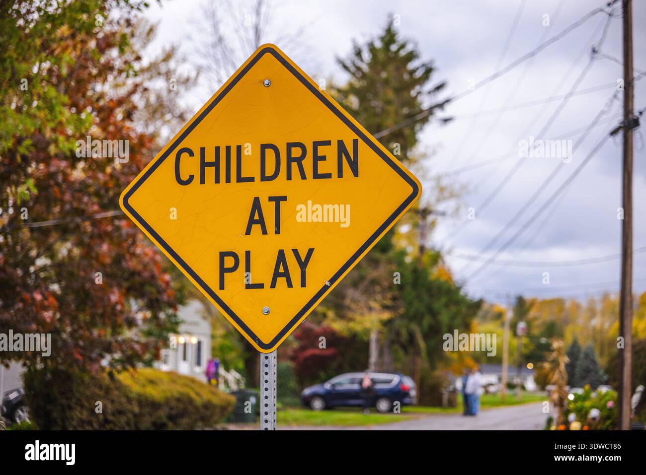 Gelbes Straßenschild mit der Aufschrift „Kinder im Spiel“ warnt Fahrer in einem Wohngebiet, umgeben von Bäumen und Häusern im Herbst. Stockfoto