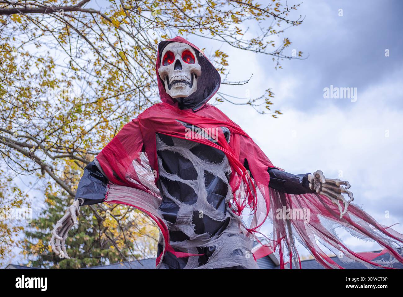 Gruselige Halloween Skelett Dekoration mit leuchtenden roten Augen und gerissenem rotem Mantel unter bewölktem Herbsthimmel. Stockfoto