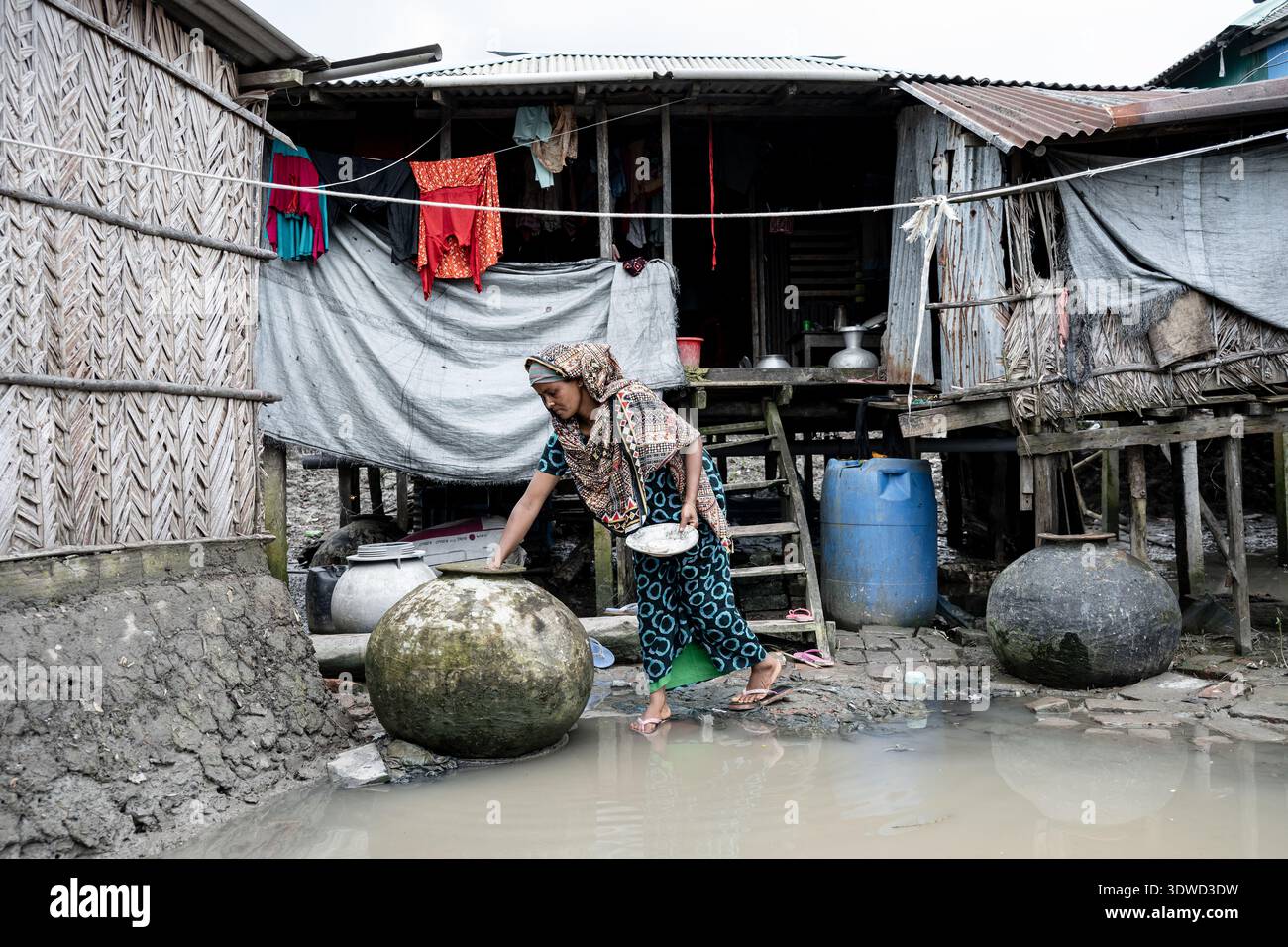 Satkhira, Bangladesch - 12. Juli 2025: Blick auf eine Frau, die auf dem überfluteten Boden in der Nähe von Pfahlhäusern navigiert, wobei die gedämpften Töne des Wassers die Überdecke reflektieren Stockfoto