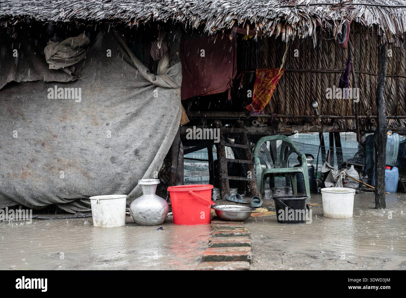 Satkhira, Bangladesch - 14. Juli 2025: Blick auf ein gestelztes Haus inmitten des unbarmherzigen Regenschusses des Monsuns, wo leuchtende rote und silberne Eimer stehen Stockfoto