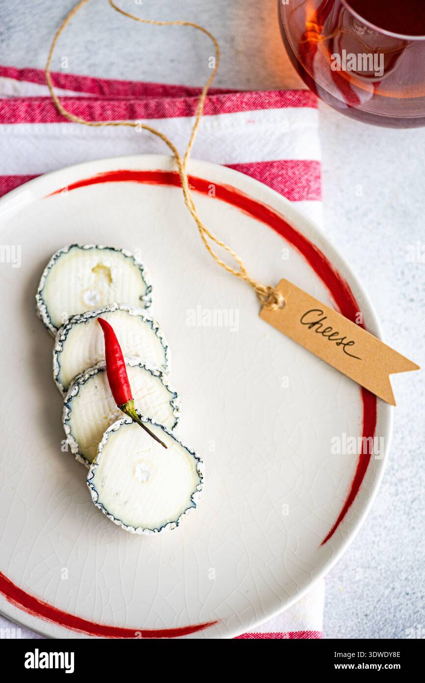 Scheiben cremigen Brie-Käses, elegant auf einem Teller angeordnet, kombiniert mit einem Glas Bernstein, auf einem rot-weißen Tuch, das einen perfekten Gour verleiht Stockfoto