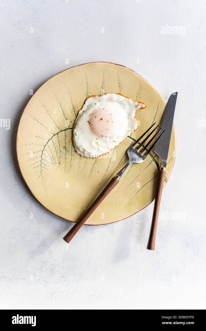 Ein perfektes Spiegelei auf einer dekorativen Keramikplatte mit Gabel und Messer, das eine einfache, aber elegante Mahlzeitenpräsentation präsentiert, ideal zum Frühstück Stockfoto