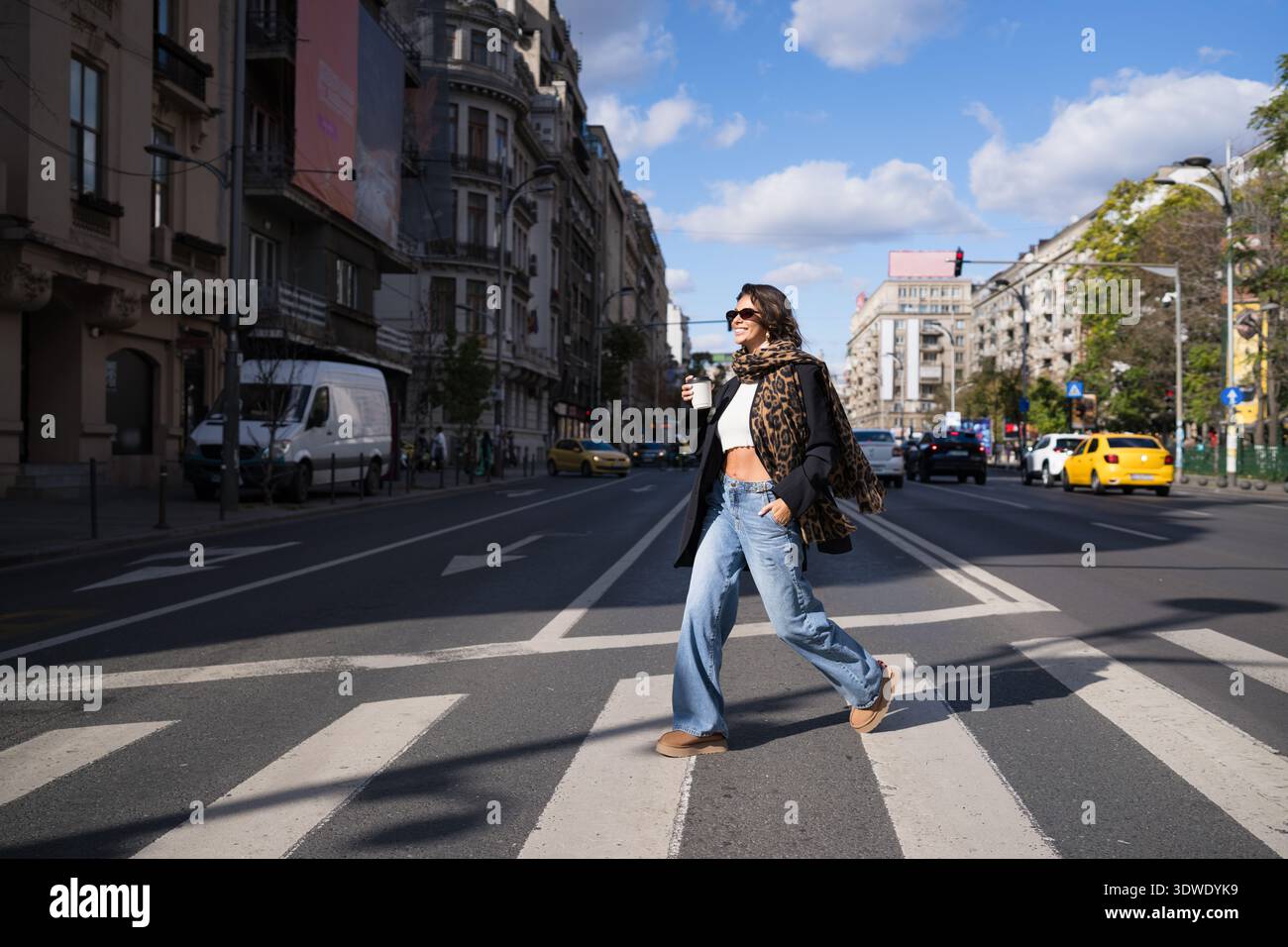 Eine stilvolle Frau mit Sonnenbrille und einem Schal mit Leopardenmuster geht lässig über eine Stadtstraße und hält eine Tasse Kaffee vor einem urbanen Hintergrund Stockfoto