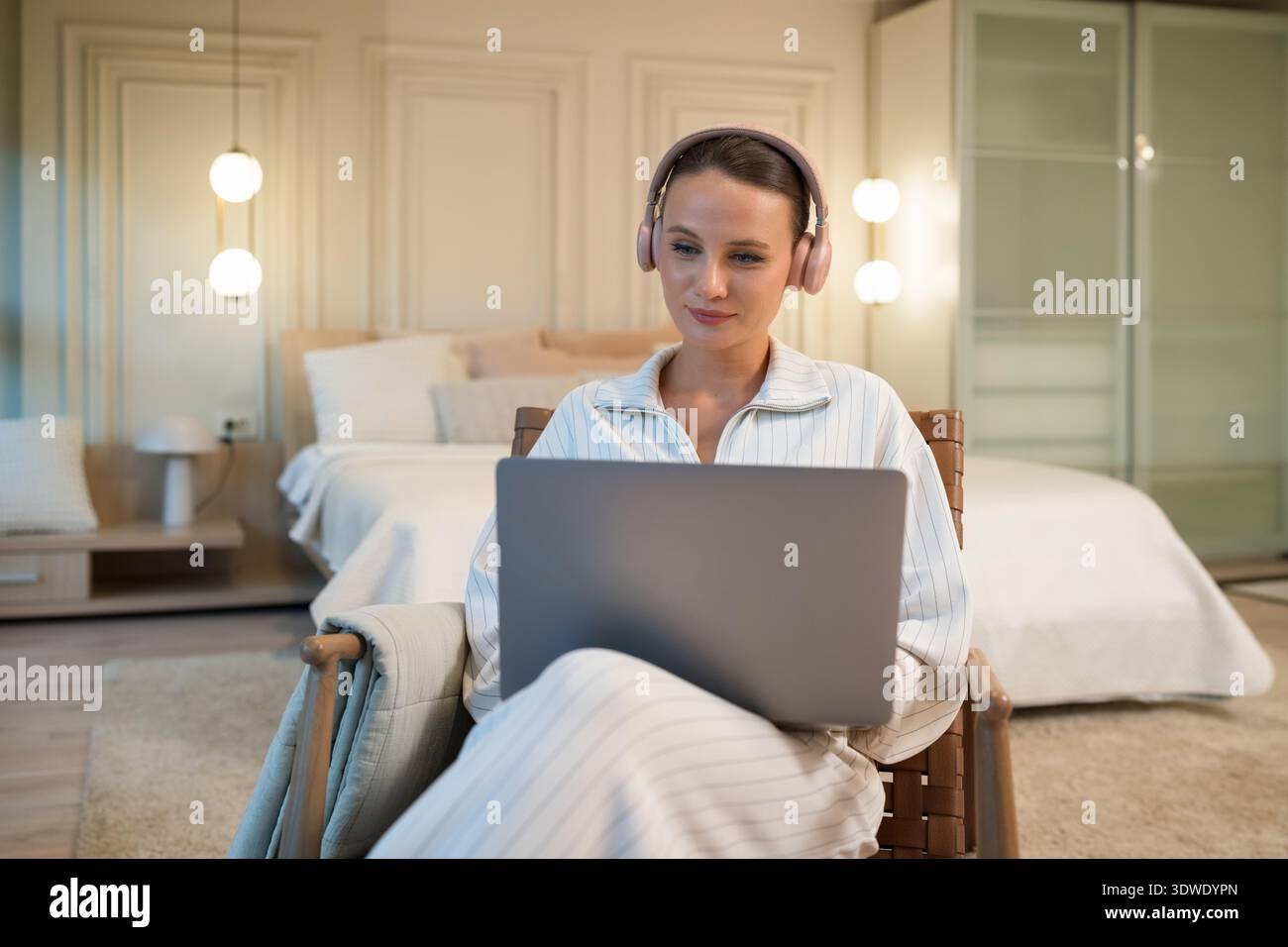 Eine junge Frau sitzt bequem in ihrem Schlafzimmer, trägt Kopfhörer und konzentriert sich auf ihren Laptop-Bildschirm. Sanfte Beleuchtung und gemütliche Einrichtung sorgen für eine warme Atmosphäre Stockfoto