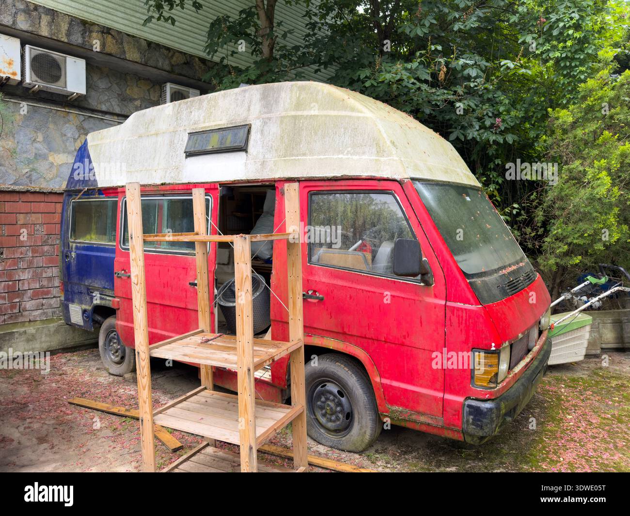 Ein verwitterter und verlassener Vintage-Van sitzt bewegungslos, seine Farbe verblasste und die Struktur zeigt Anzeichen von Rost und Verfall. Umgeben von Grün, erzählt es Stockfoto