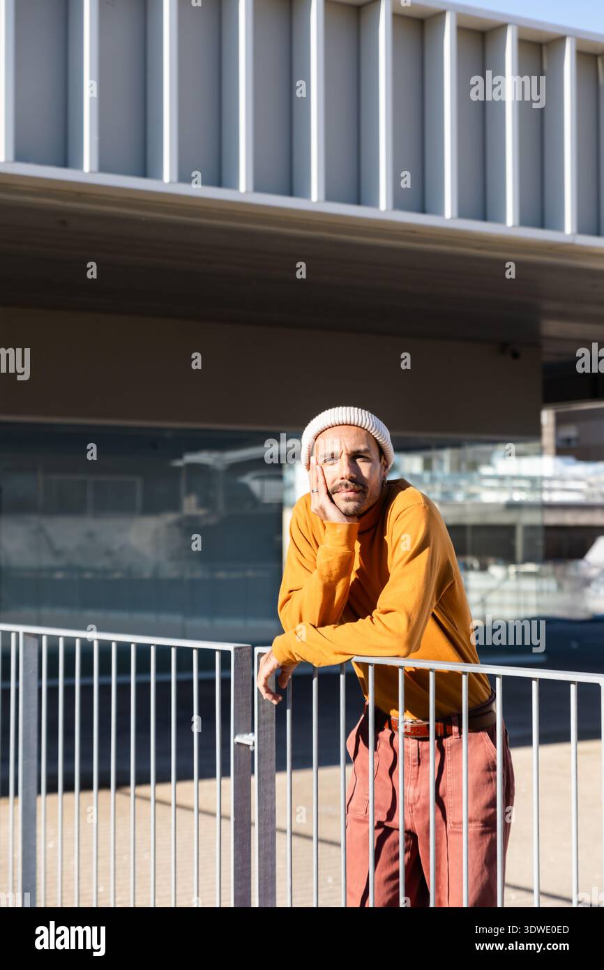 Ein Mann lehnt sich lässig auf ein Metallgeländer im Freien, trägt einen orangefarbenen Pullover und eine weiße Mütze. Die entspannte Pose vermittelt ein Gefühl von conte Stockfoto