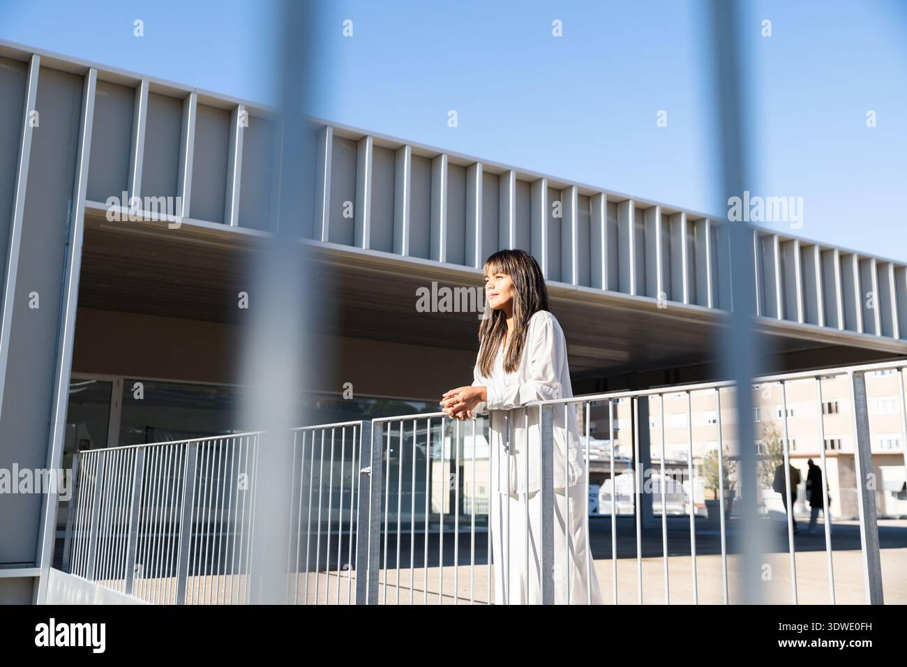 Eine Frau steht draußen und lehnt sich auf ein Geländer mit einem ruhigen Ausdruck. Das Ambiente bietet moderne Architektur und helles Tageslicht. Ihr entspannter Körper Stockfoto