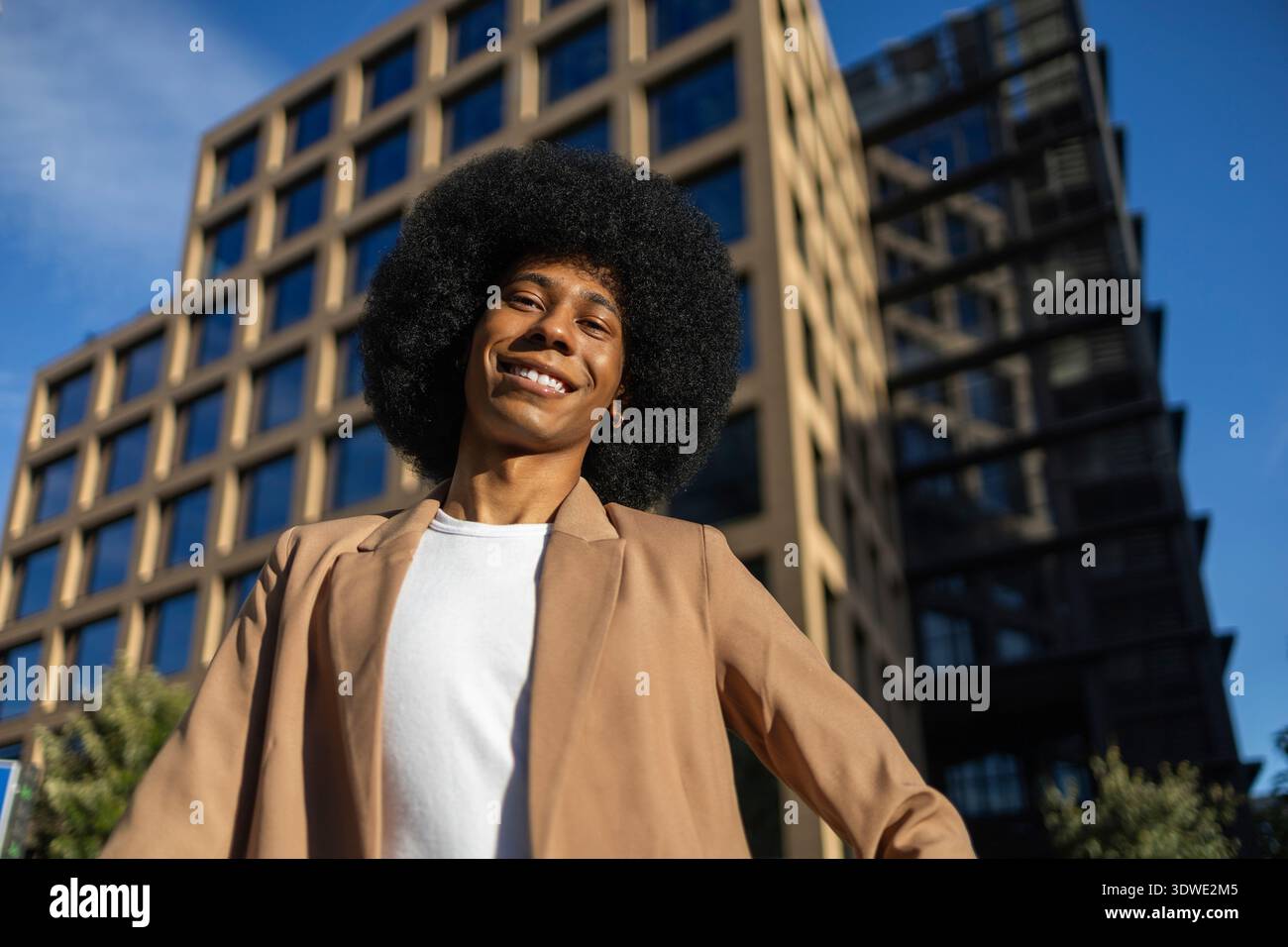 Eine Person mit braunem Blazer lächelt selbstbewusst vor der Kulisse eines modernen Bürogebäudes. Der klare blaue Himmel verstärkt die Lebendigkeit und den Erfolg Stockfoto