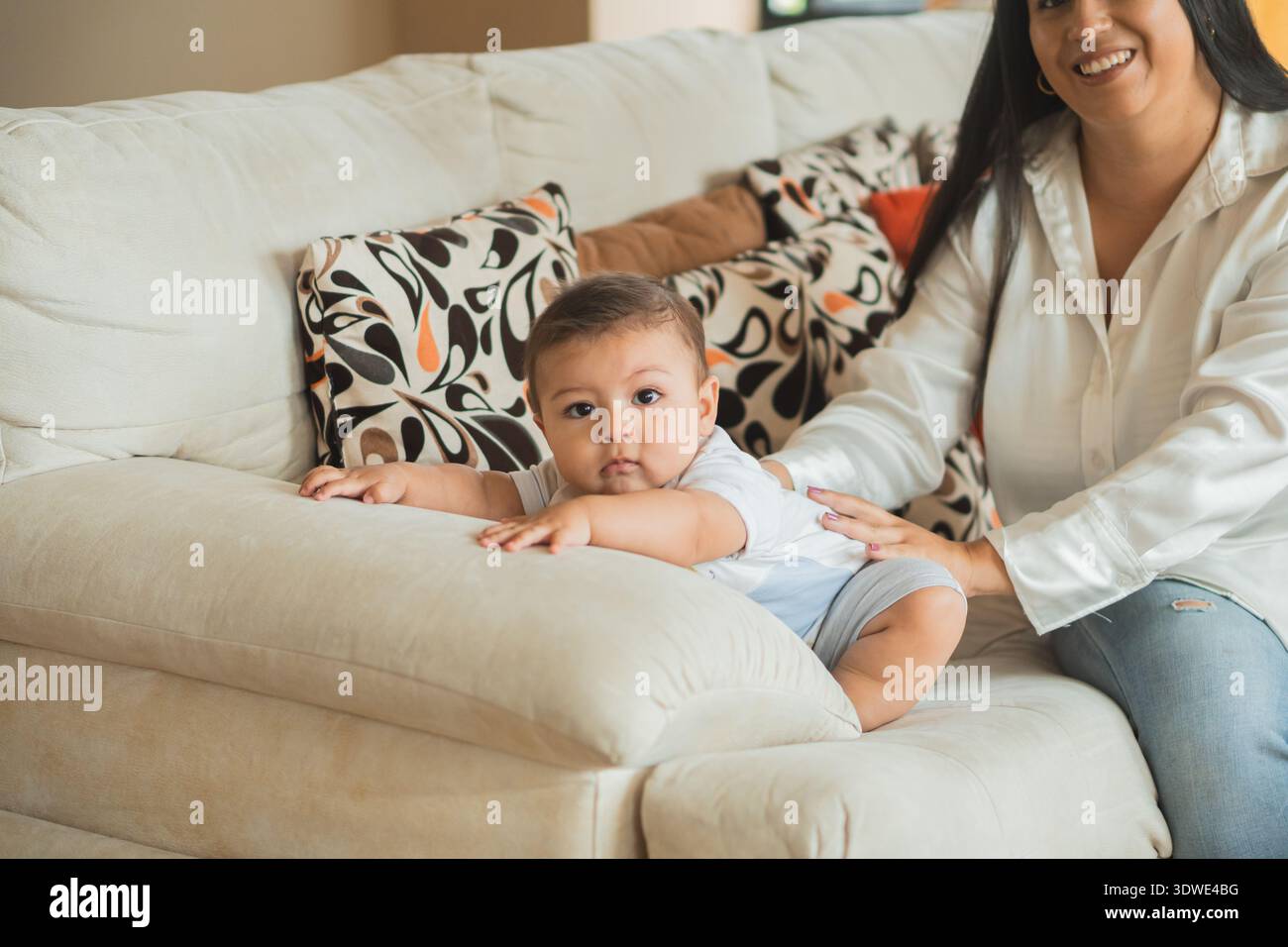 Ein entzückendes Latein-Baby liegt auf einem gemütlichen Sofa, das sanft von einem lächelnden Elternteil gestützt wird. Das Wohnzimmer verfügt über farbenfrohe, gemusterte Kissen, Crea Stockfoto