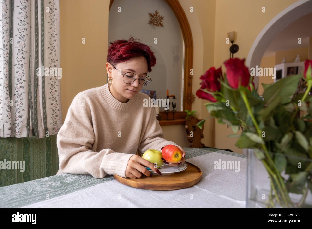 Eine junge Frau mit Brille und rotem Haar sitzt an einem Esstisch und genießt einen Moment zu Hause. Sie trägt einen kuscheligen Pullover und ist von einem warmen, invit umrahmt Stockfoto