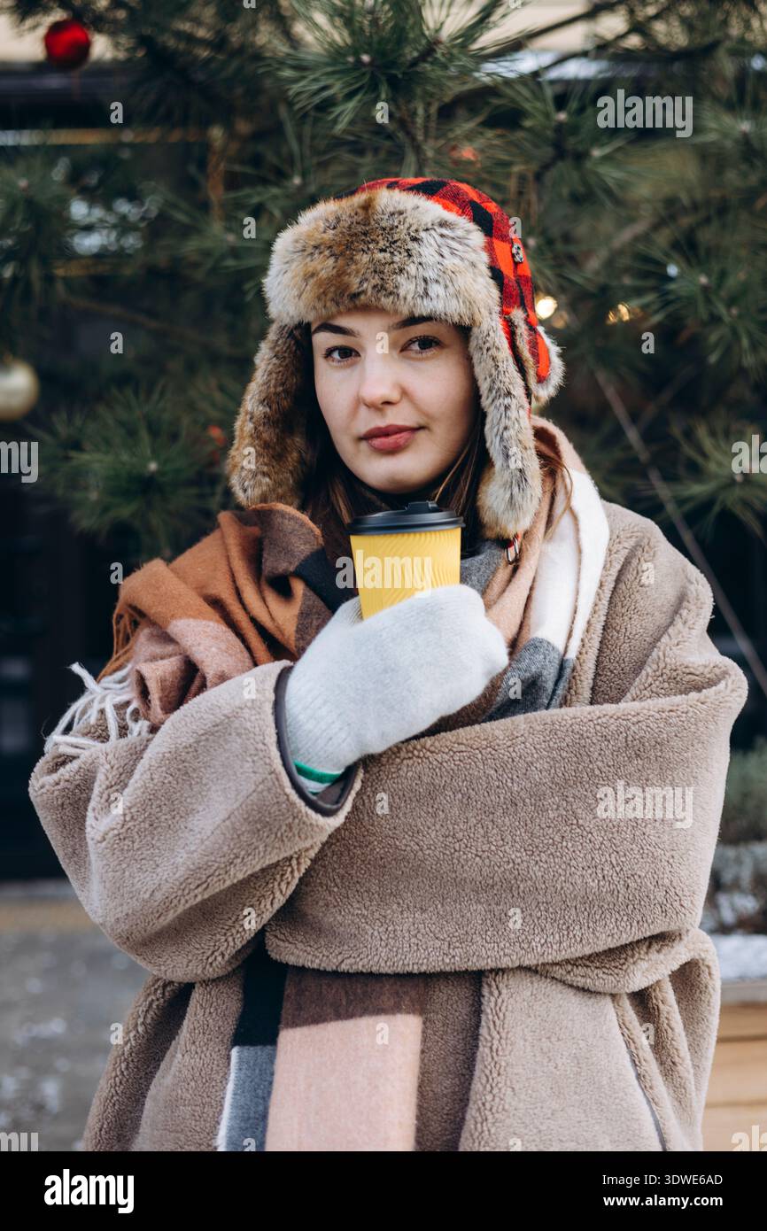 Eine Frau in einen gemütlichen Wintermantel gewickelt hält draußen eine heiße Kaffeetasse. Sie trägt einen Hut und Fäustlinge mit Fellfutter, die die Wintersaison mit Wärme umhüllen Stockfoto