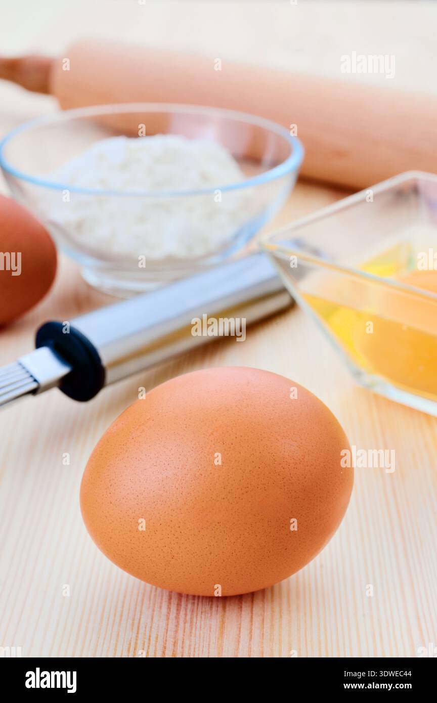 Ein Nahaufnahme-Bild, das wichtige Backzutaten zeigt, darunter ein braunes Ei, eine Schüssel Mehl und eine kleine Schüssel mit zerbrochenen Eiern auf einem Holztisch, Stockfoto