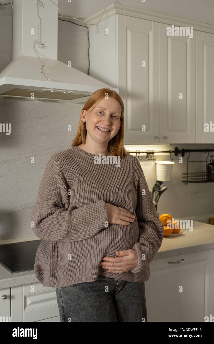 Eine schwangere Frau steht lächelnd in einer gemütlichen Küche und hüllt sanft ihren Bauch. Sie wirkt zufrieden und entspannt, umgeben von ihrer warmen Atmosphäre Stockfoto