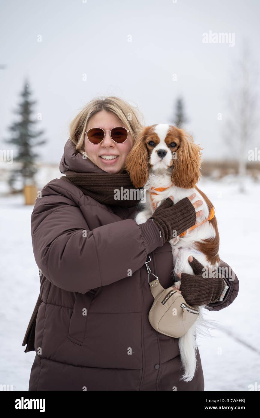 Eine Frau in Wintermantel und Schal hält ihren Kavalier König Charles Spaniel. Sie sind draußen an einem schneebedeckten Tag und verkörpern Wärme und Komfort Stockfoto